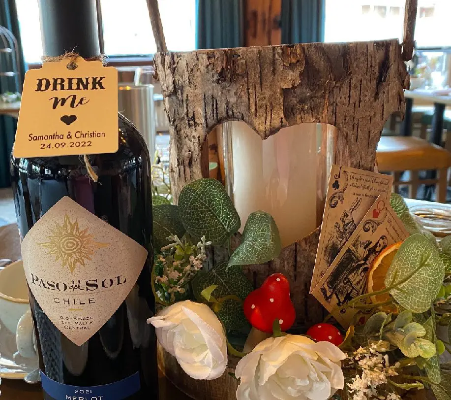 A wine bottle labeled "Paso del Sol" sits beside a rustic wooden candle holder adorned with flowers and small cards. Perfect for a charming wedding, the tag reads "Drink Me" alongside names and dates, capturing the essence of personalized celebration planning.