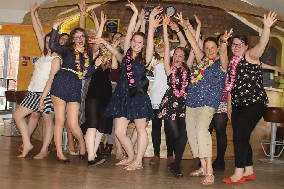 A lively group of people celebrating indoors, adorned with colorful leis and some with arms raised, perfectly captures the joyous spirit of a wedding. One person, wearing a "Bride to Be" sash, hints at careful planning that makes this moment unforgettable.