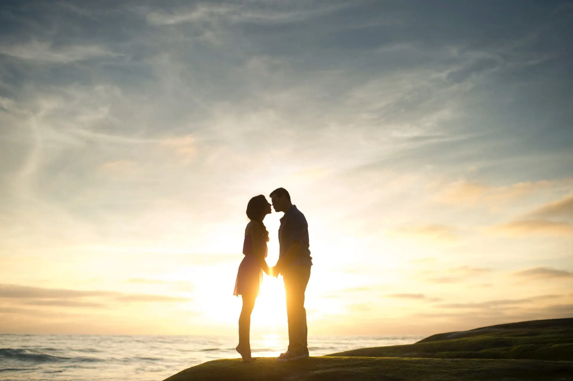 Silhouette of a couple standing close on a hill, with the sun setting over the ocean in the background, creating a perfect moment for a wedding business to capture and cherish.