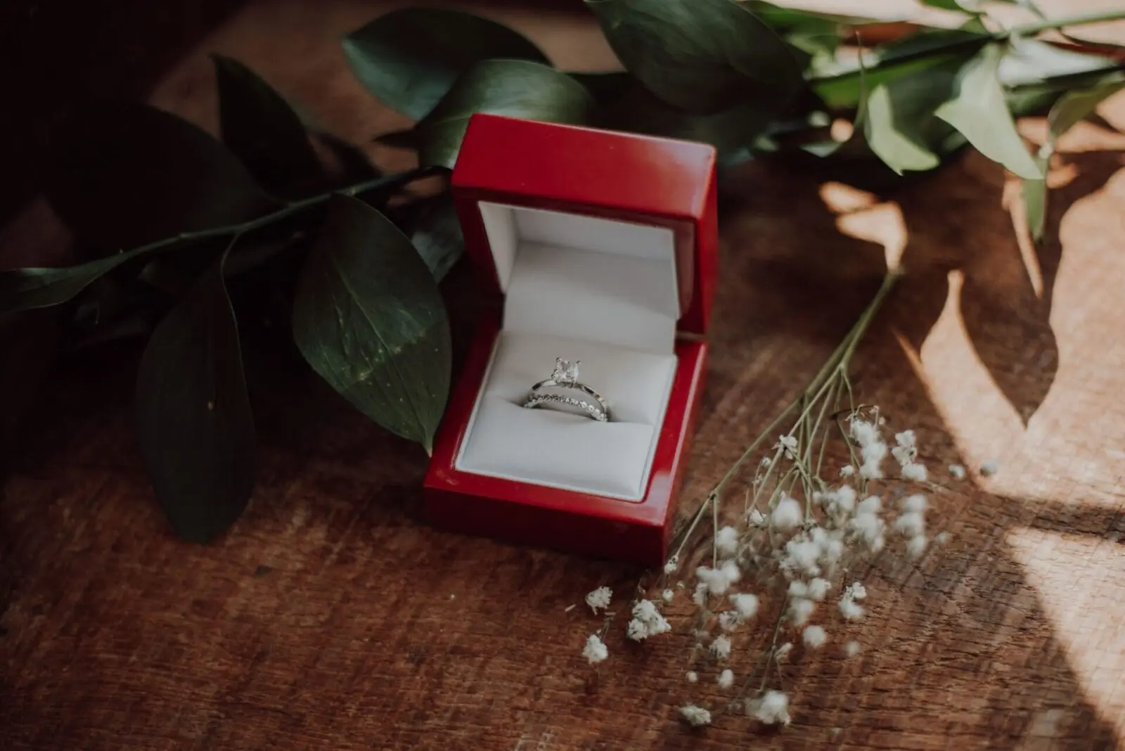 An engagement ring set in a red jewelry box, surrounded by green leaves and white flowers, rests on a wooden surface—an ideal inspiration for those planning their wedding.
