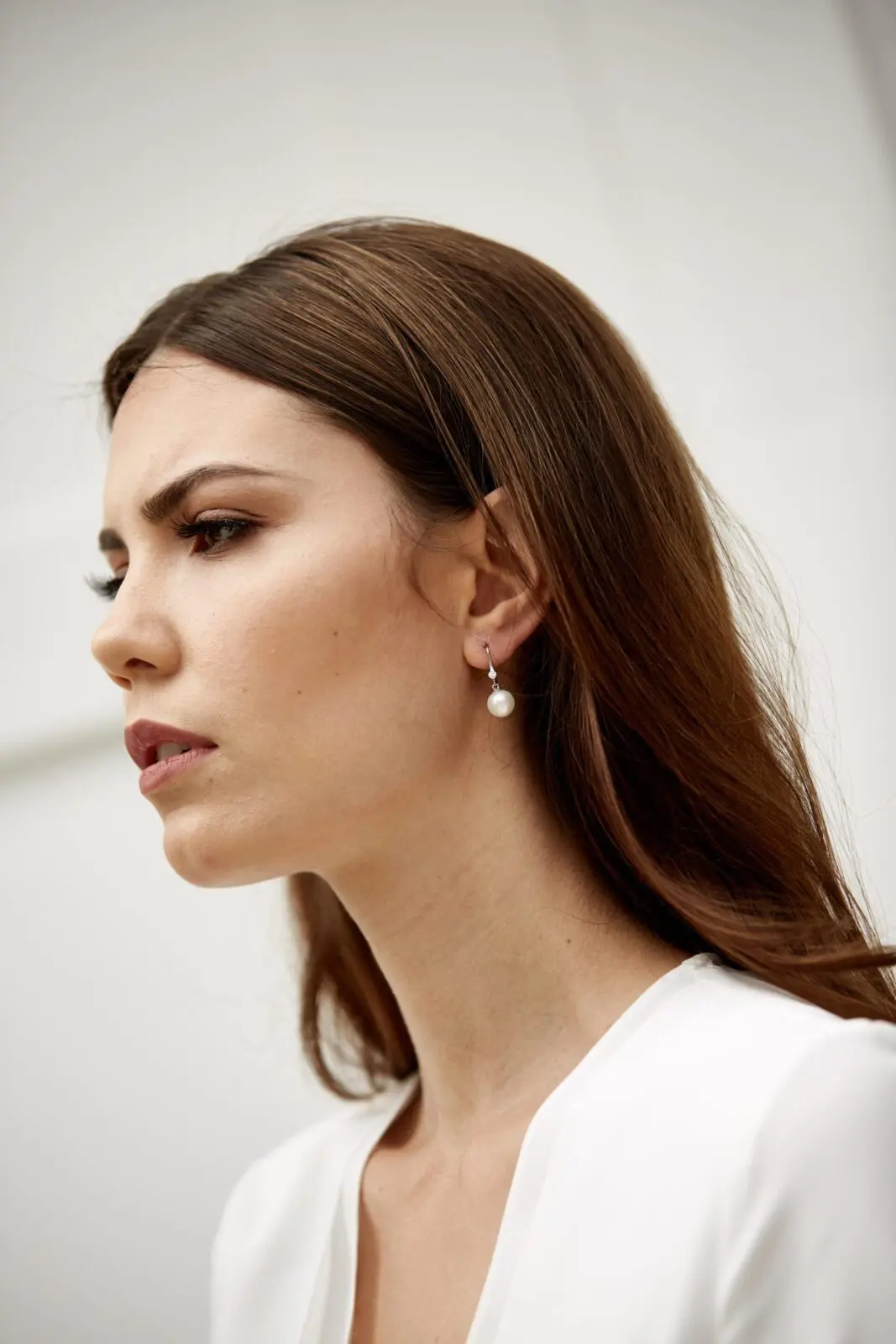 Side profile of a person with long brown hair and pearl earrings, wearing a white top, looking contemplative, perhaps reflecting on the joys of a wedding.
