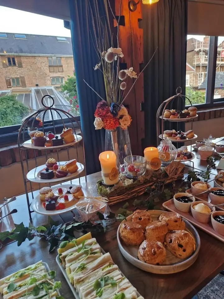 The table is elegantly set for afternoon tea, featuring sandwiches, scones, and desserts, with the gentle glow of candles and a floral centerpiece. Perfect for a wedding planning showcase, it offers a picturesque view of the buildings outside.