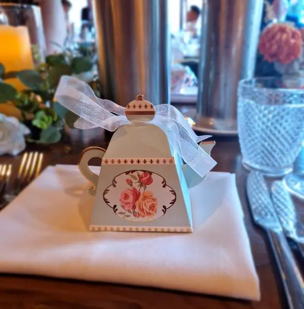 A decorative teapot-shaped box with a floral design and white ribbon sits on a napkin, surrounded by table settings and a floral centerpiece, adding charm to an elegant wedding atmosphere.