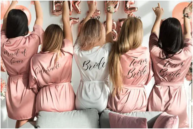 Five women in robes sit on a sofa facing away, showing the words: "Maid of Honor," "Bridesmaid," "Bride," and "Team Bride." Pink and white are the robe colors, adding a charming touch to this moment in wedding planning.