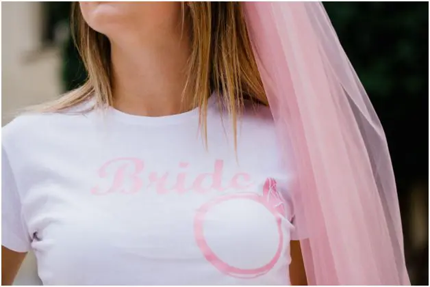A person wearing a white T-shirt adorned with the word "Bride" and a ring design, perfectly paired with a pink veil—a delightful touch in wedding planning.