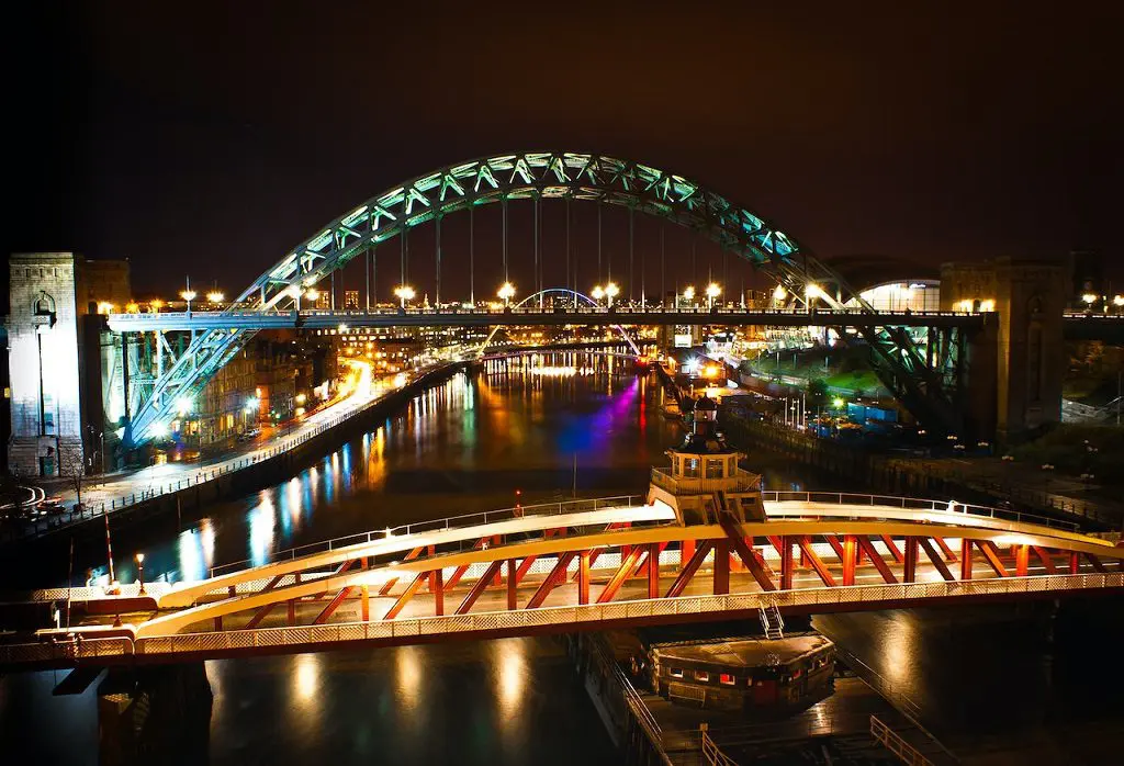 A night view of a lit arched bridge over a river, with reflections shimmering on the water, surrounded by other illuminated structures—an enchanting scene perfect for a wedding planner seeking the ideal romantic backdrop in the UK.