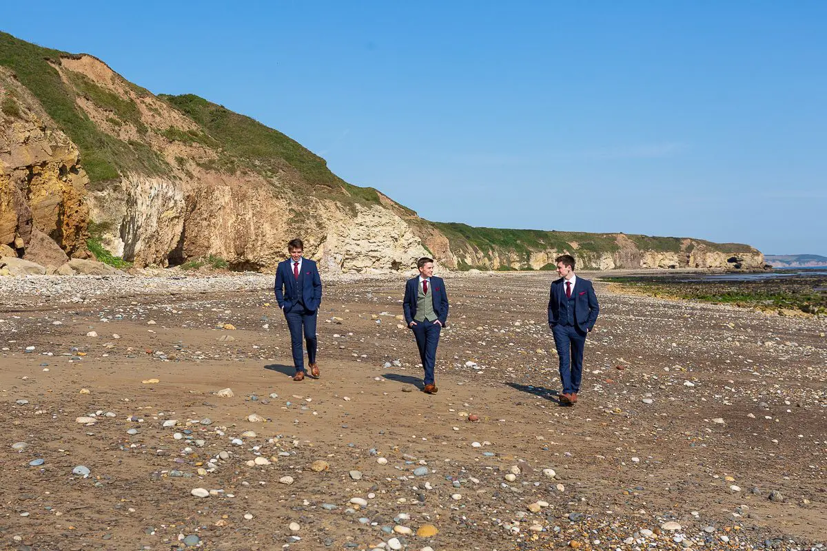 Three people in suits walk along a rocky beach with cliffs in the background under a clear blue sky, perhaps brainstorming ideas for a unique wedding business or setting plans for a stunning wedding in the UK.