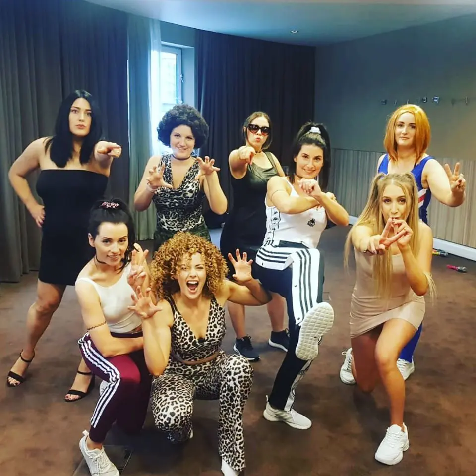 Eight women in various poses and outfits, including animal prints, tracksuits, and athletic wear, discuss wedding planning in a room with curtain backdrop.