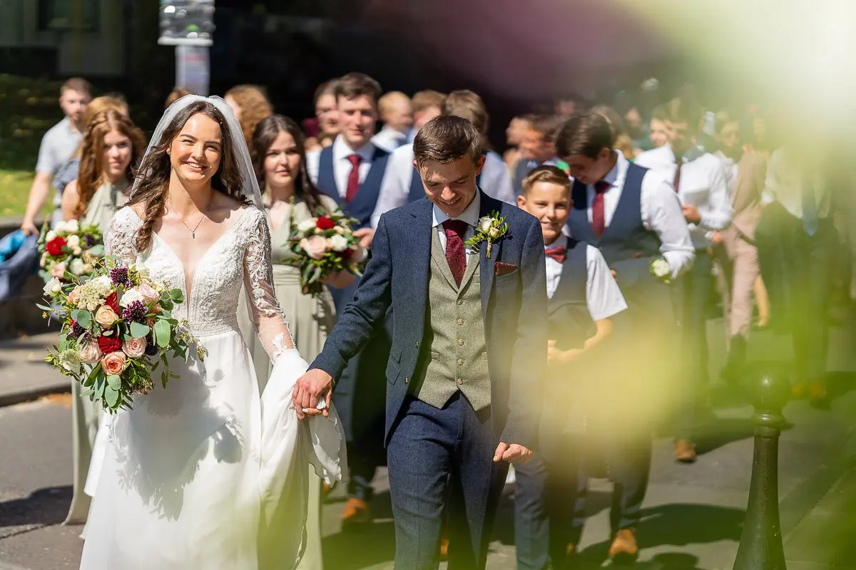 A bride and groom walk hand in hand, immersed in a well-planned celebration with their wedding party. The bride, smiling brightly, holds a bouquet of flowers.