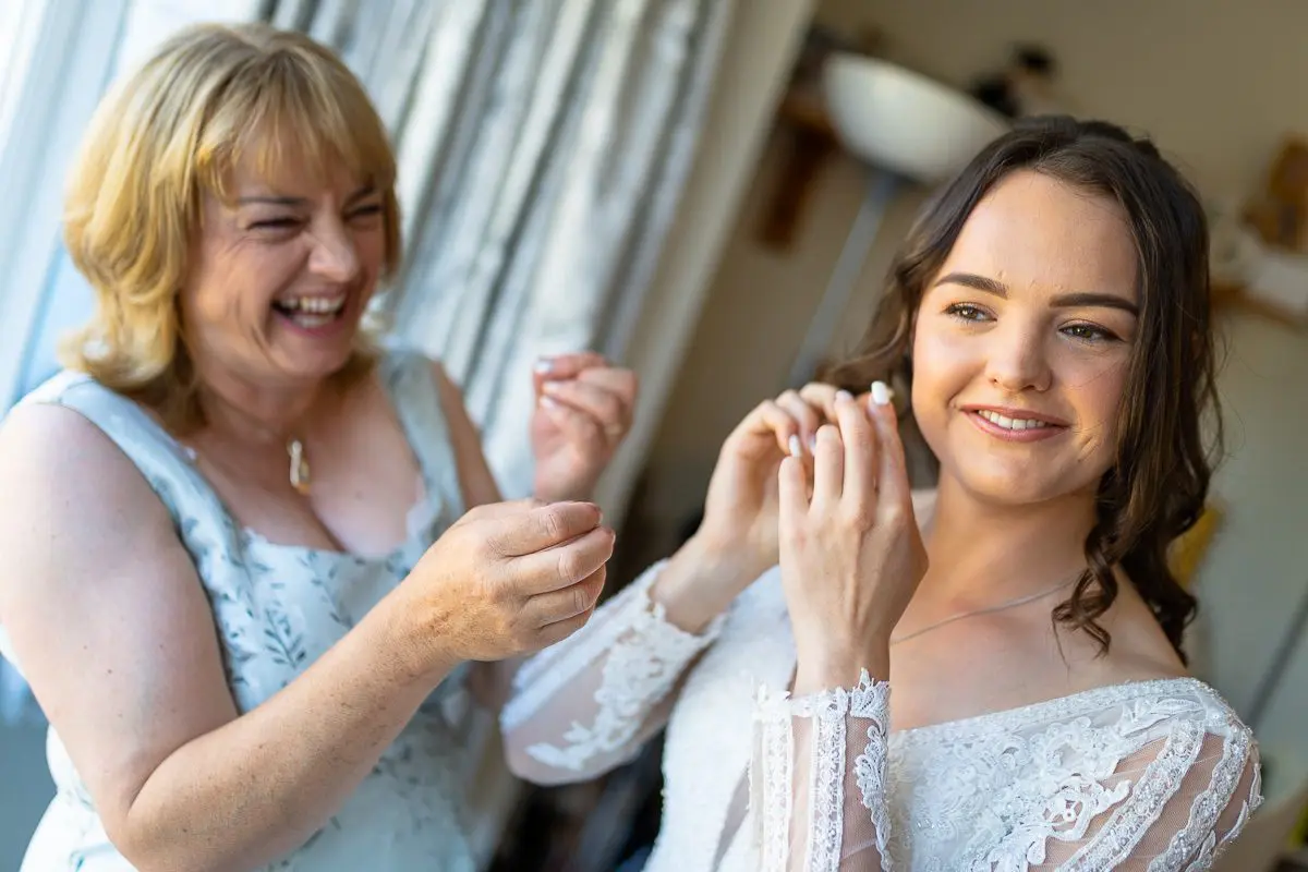 A woman helps another adjust her earrings, both grinning by the window. The woman on the right, in a white lace dress, radiates joy as part of the delightful wedding preparations.