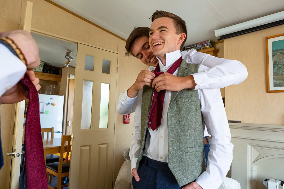 In a cozy room where planning meets elegance, one person helps the other adjust a tie. Their smiles are radiant, reflecting the joy of wedding services. Dressed in formal attire, they stand with a door and dining area gently framing their special moment.