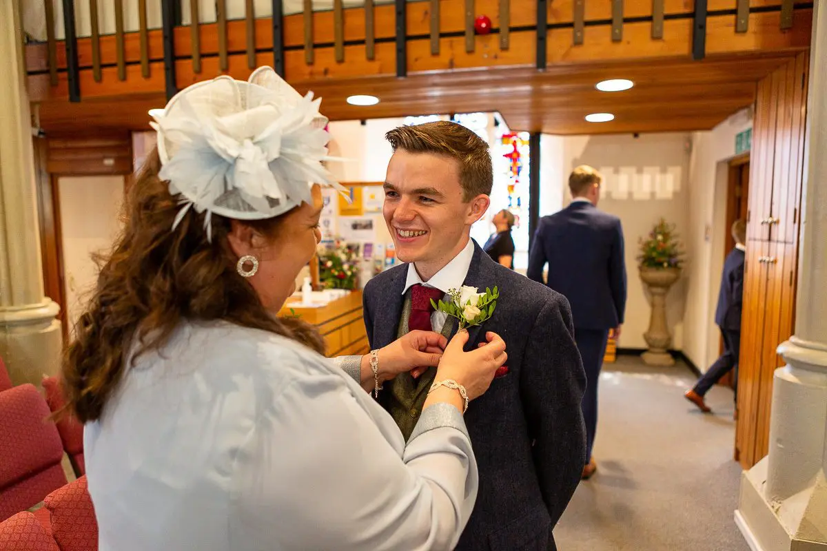 In a warmly decorated room, a woman adjusts a flower on a young man's suit as part of wedding planning. Both are smiling.