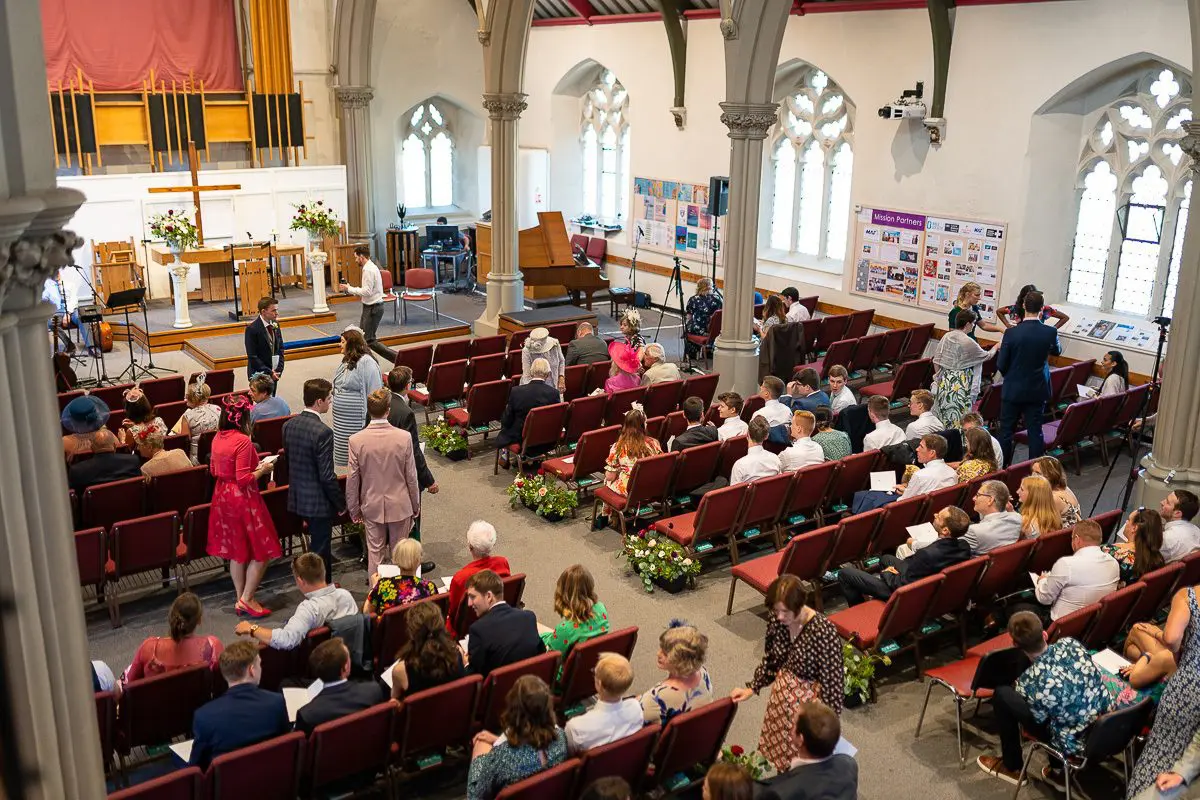 A diverse group of people seated in a church with tall windows, preparing for a wedding. Floral decorations and a cross are visible at the front, setting the perfect scene for this joyful UK event.