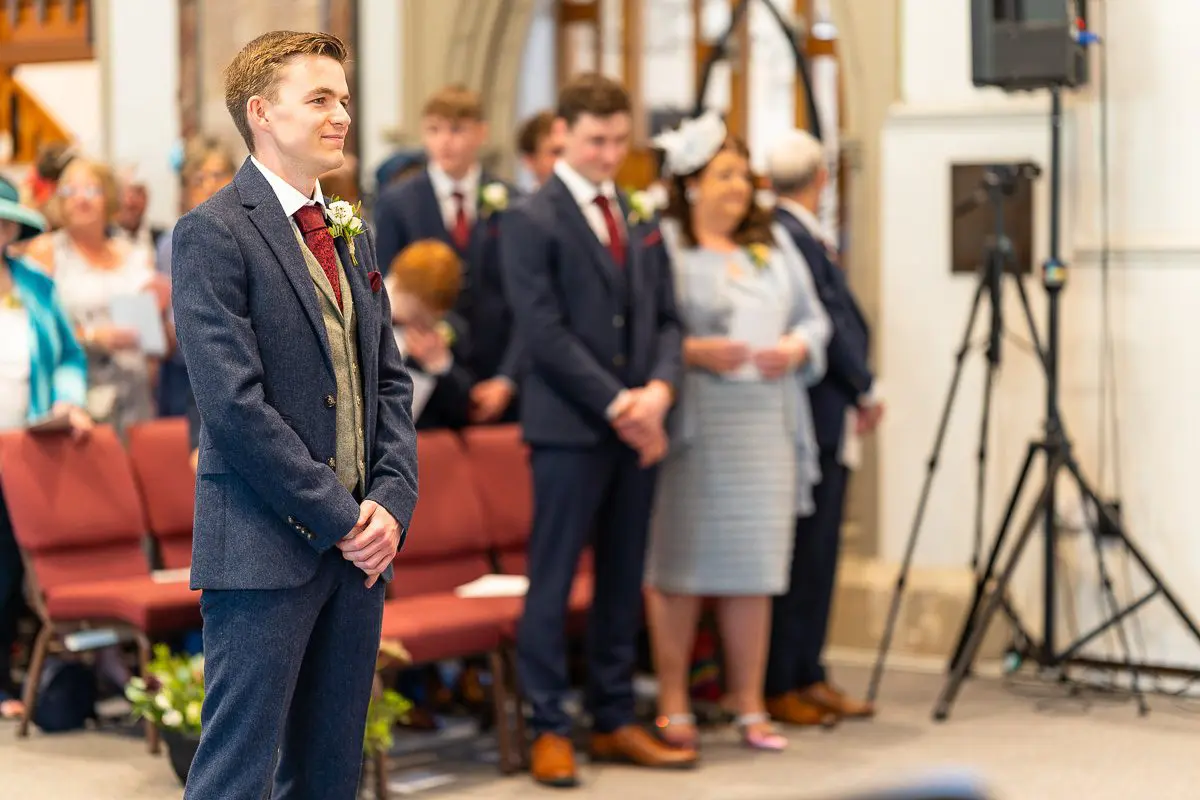 A groom stands in a church, smiling, as guests from across the UK gather behind him. He wears a dark suit with a red tie, embodying elegance. A camera is set up in the background for capturing moments provided by top-tier wedding services.