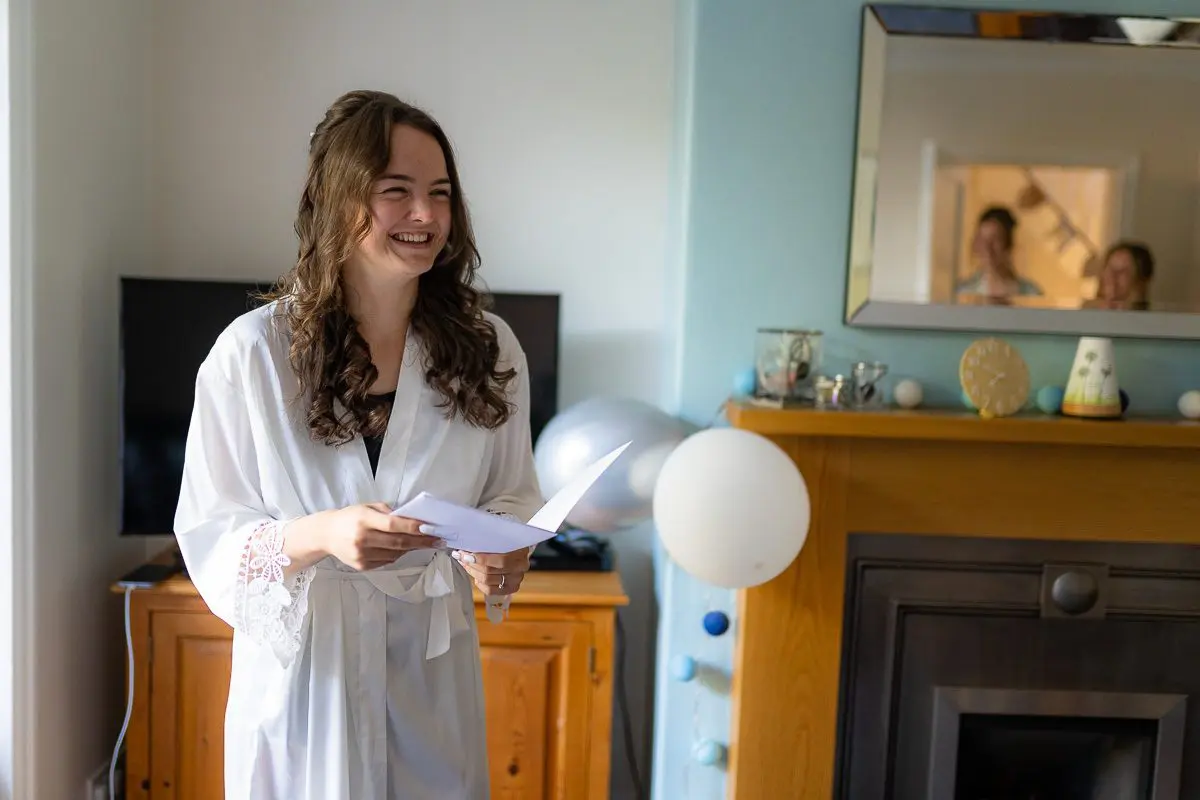 A smiling woman in a white robe, immersed in wedding planning, holds papers in a room adorned with a mirror, TV, and decorative balloons.