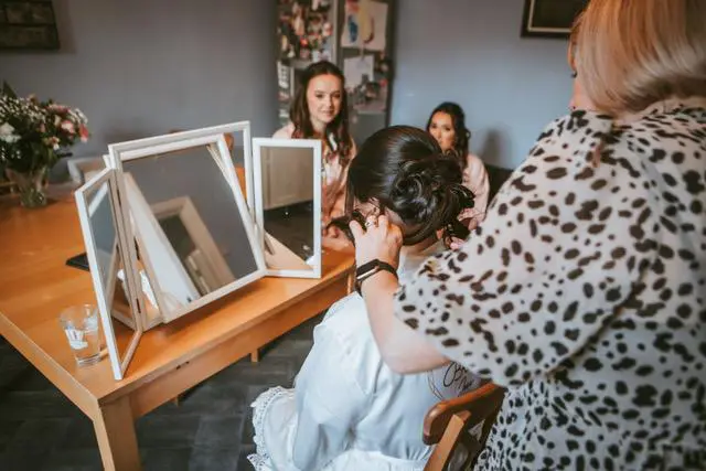 A woman expertly styles another woman's hair at a table with a three-panel mirror, capturing the essence of wedding planning. In the background, two observers are seated, watching attentively. The room is beautifully adorned with decorations and flowers, enhancing the scene's charm.