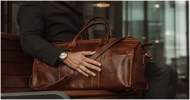 A person sits on a bench in a sharp suit and elegant watch, looking through a large brown leather bag, perhaps finalizing their wedding planning essentials.