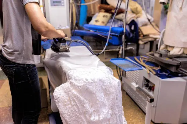 In a laundry room filled with equipment, someone is carefully ironing a white lace garment, perhaps preparing it for an elegant occasion in the bustling wedding business.