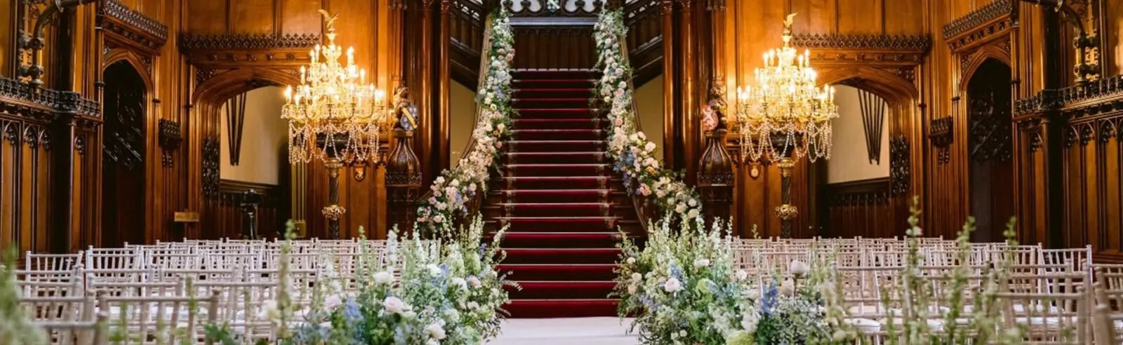 An ornate indoor space perfect for a wedding, featuring a grand staircase adorned with floral arrangements. Rows of white chairs are elegantly arranged for the event, while two large chandeliers cast a warm glow from above. Ideal for any wedding business in the UK.