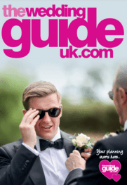 Two men in suits, one wearing sunglasses, have a corsage pinned on his jacket. Pink text at the top reads "theweddingguideuk.com," your ultimate resource for wedding planning. A pink heart with text adorns the bottom right.