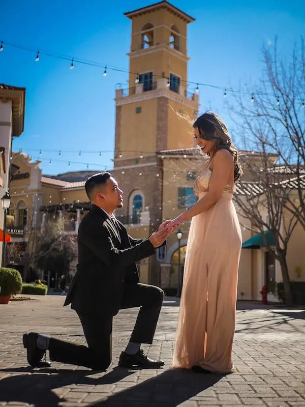 A man kneels down to propose to his girlfriend in front of a clock tower.