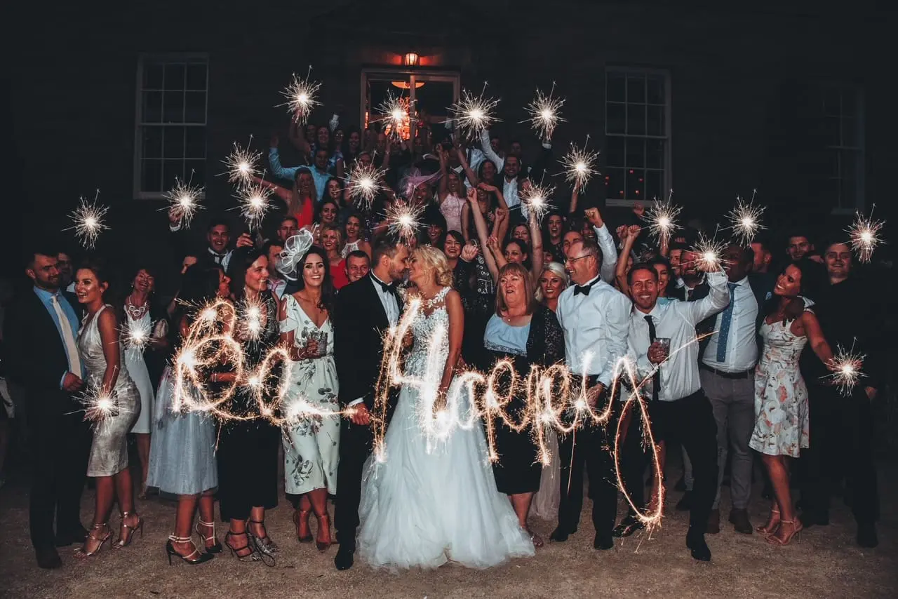 Wedding guests and newlyweds holding sparklers at night in front of a building, with "be happy" written in sparkler light.