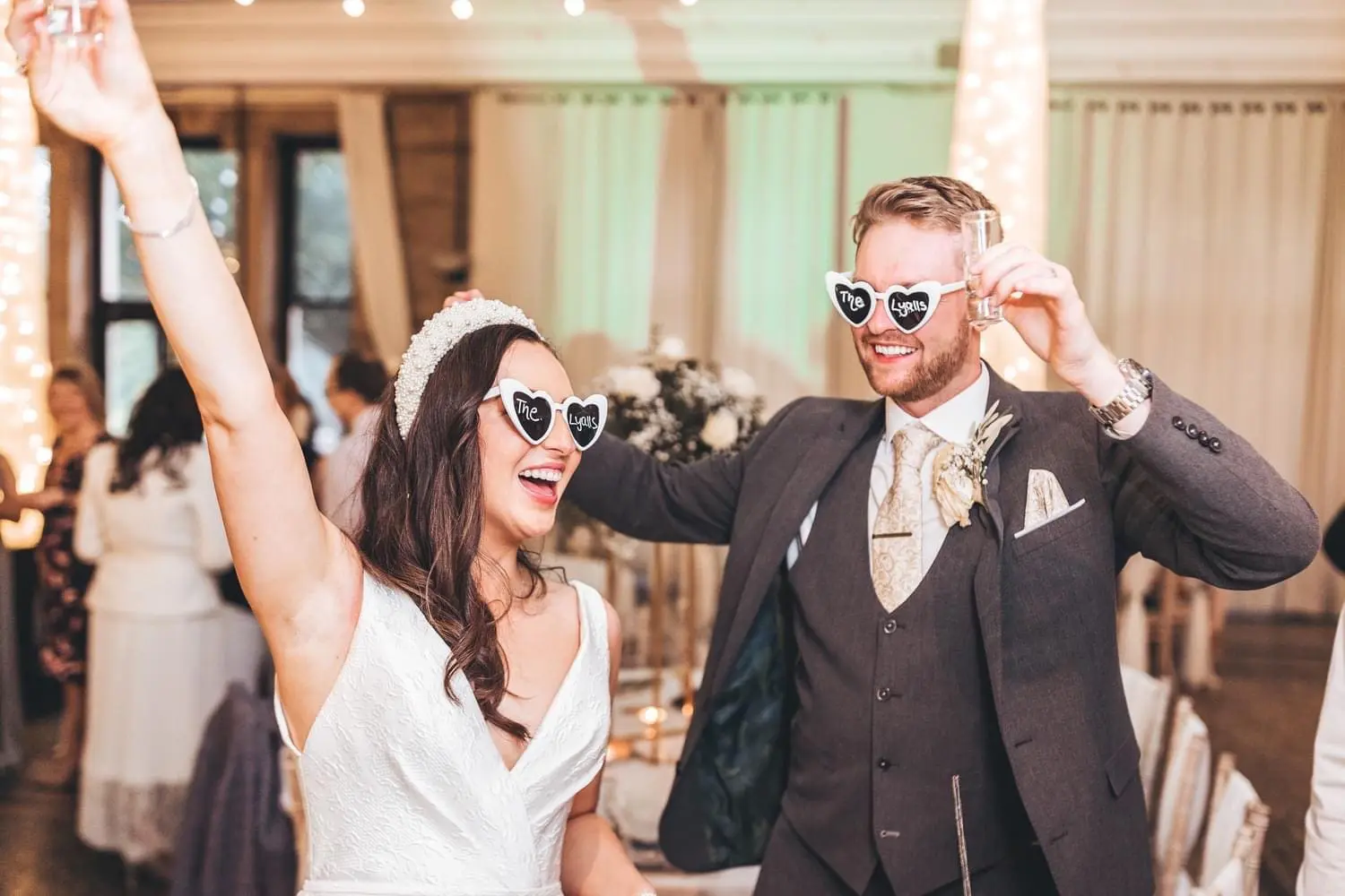A joyful bride and groom wearing heart-shaped sunglasses dance at their wedding reception, surrounded by guests.