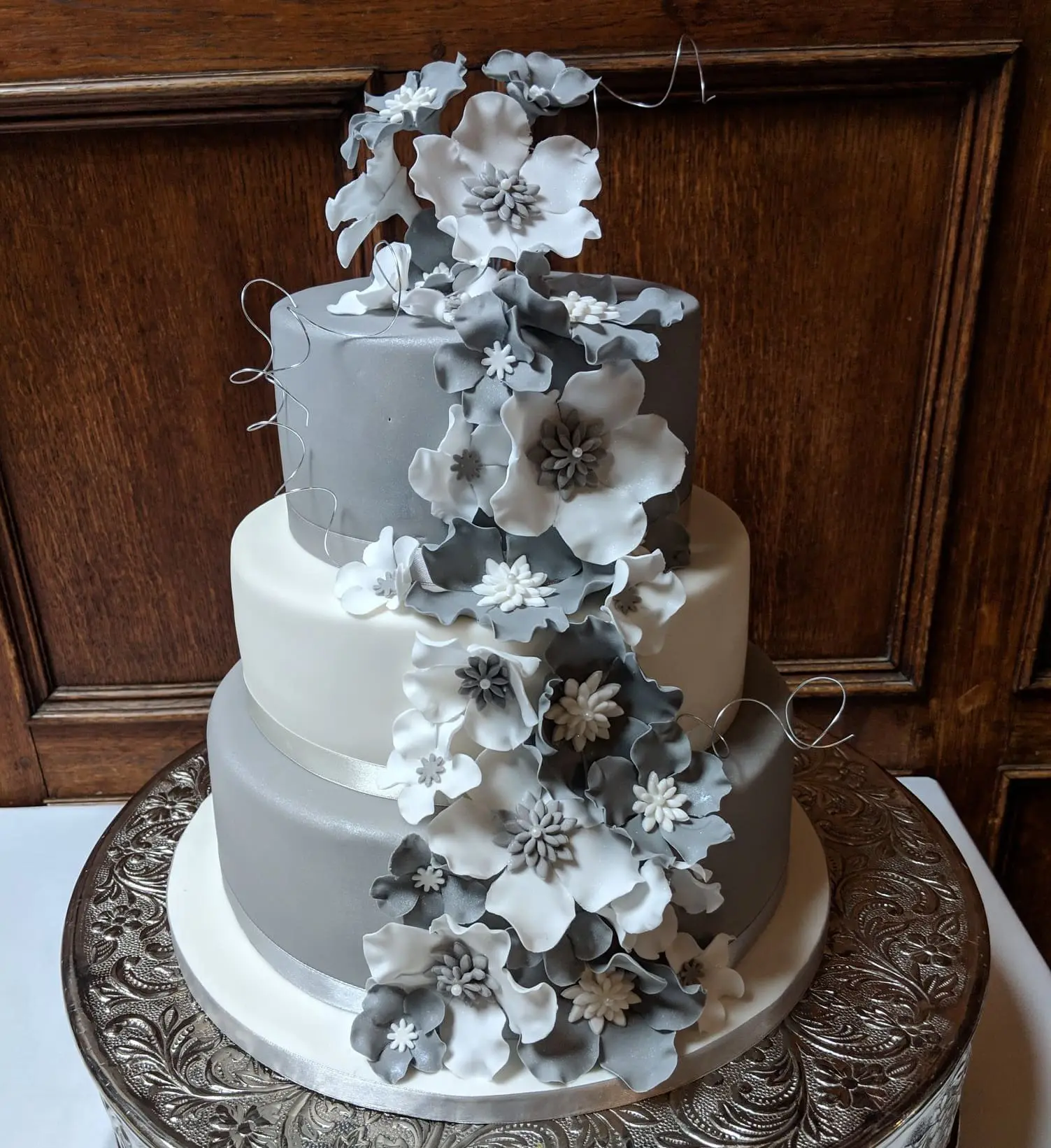A stunning three-tiered wedding cake with gray and white layers, adorned with cascading floral designs, sits elegantly on an ornate silver stand—a perfect centerpiece for any wedding celebration.