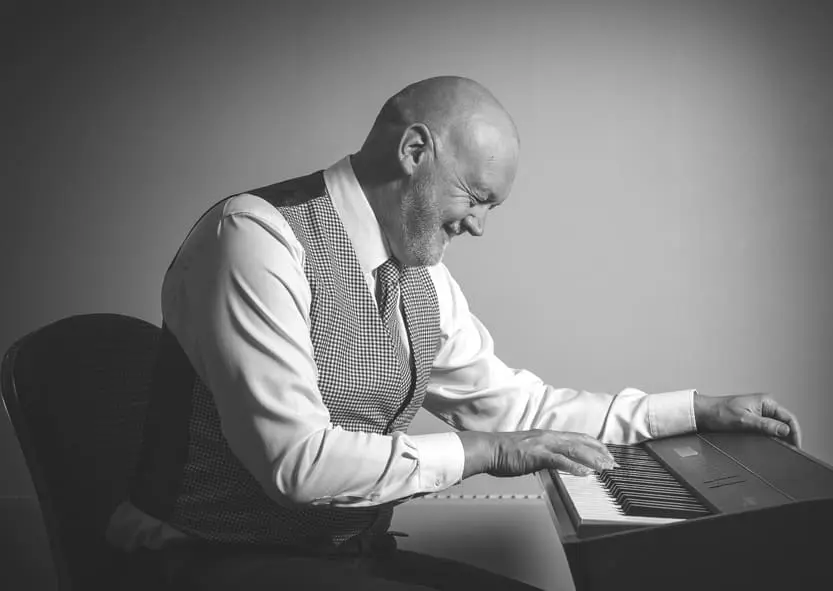 A bald man with a beard wearing a vest plays an electronic keyboard while smiling. the image is in black and white.