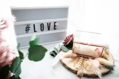 A light-themed decorative setup featuring a sign with "#love", surrounded by soft pink floral accents and a rustic wooden element.