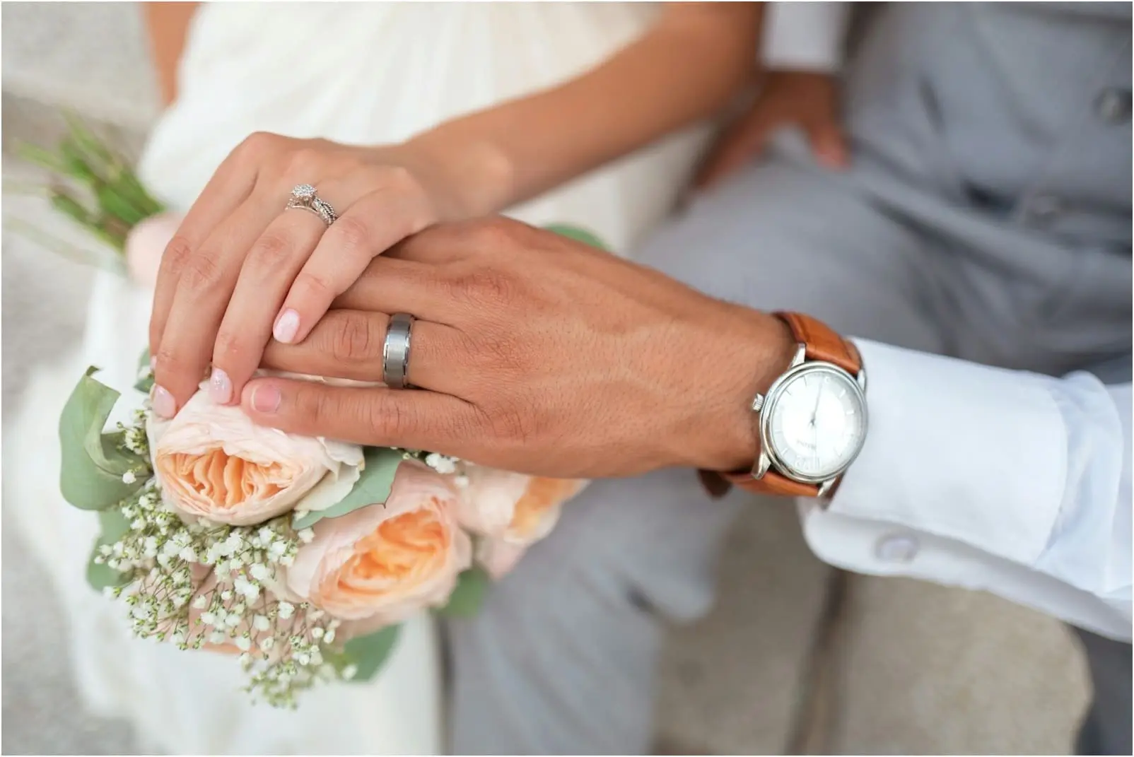 Close-up of a newlywed couple's hands showing wedding rings, the bride holding a bouquet of peach roses and baby's breath.