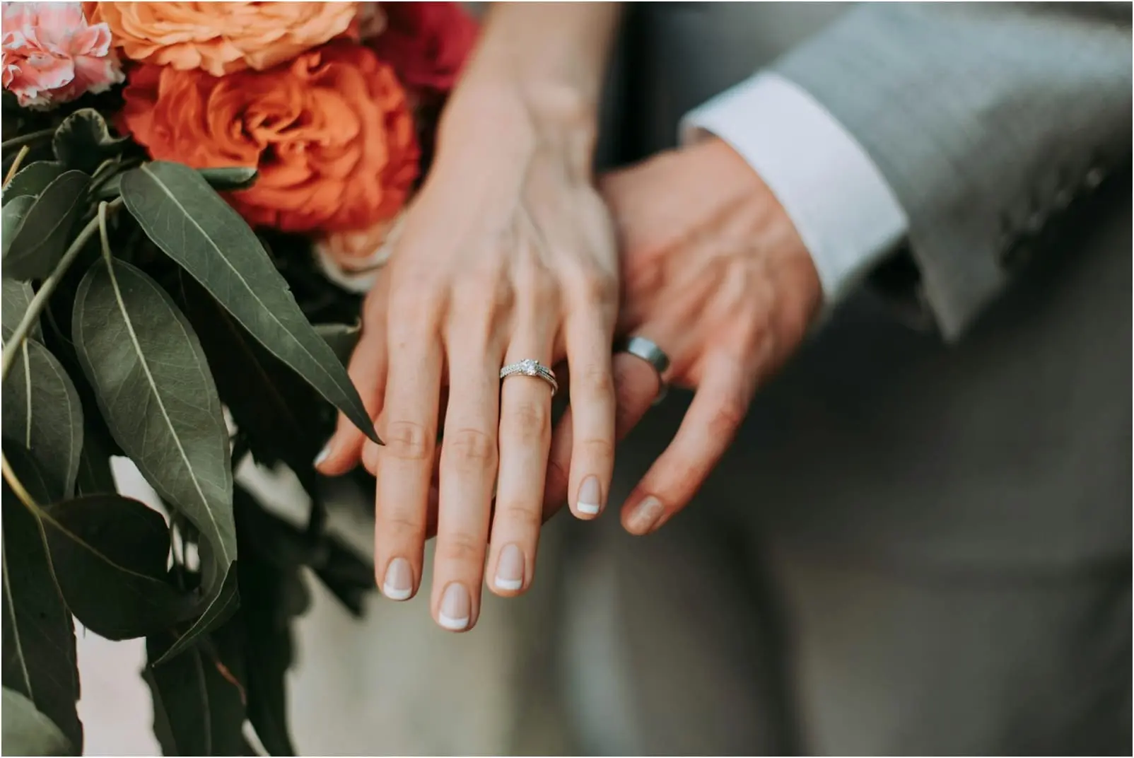 A close-up of a woman's hand with an engagement ring, resting atop a man's hand, beside a bouquet of flowers.