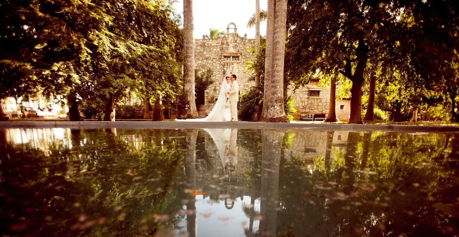 A bride and groom stand near a large reflective pond surrounded by lush greenery and tall trees, with a historic stone building in the background.