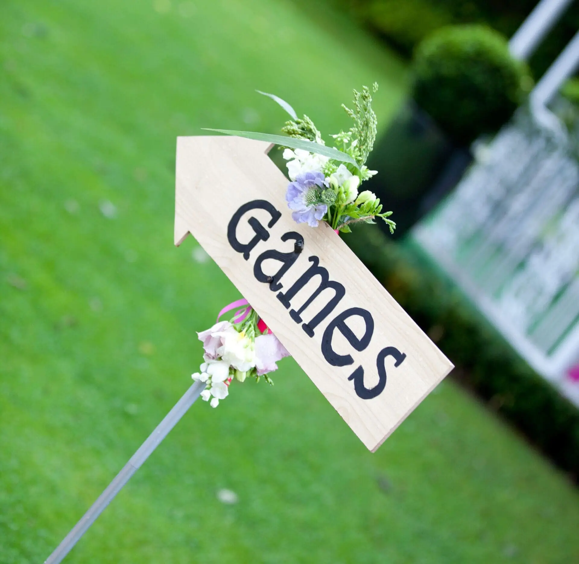 A small craft project wooden arrow sign with the word "Games" adorned with flowers, on a green lawn.