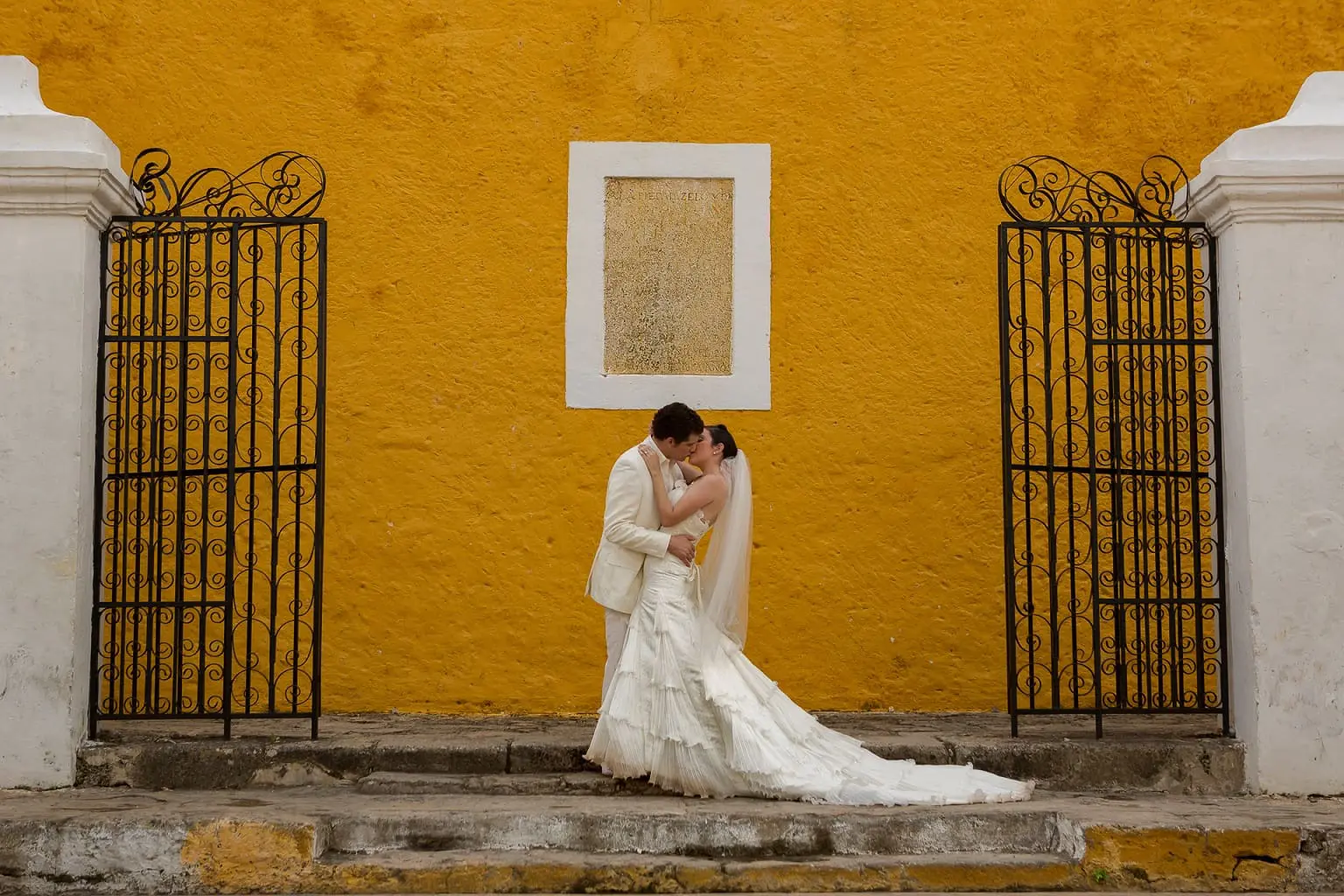 A bride and groom stand together on stone steps against a bright yellow wall with black iron gates on either side. The bride wears a white gown and veil, and the groom is in a light-colored suit.