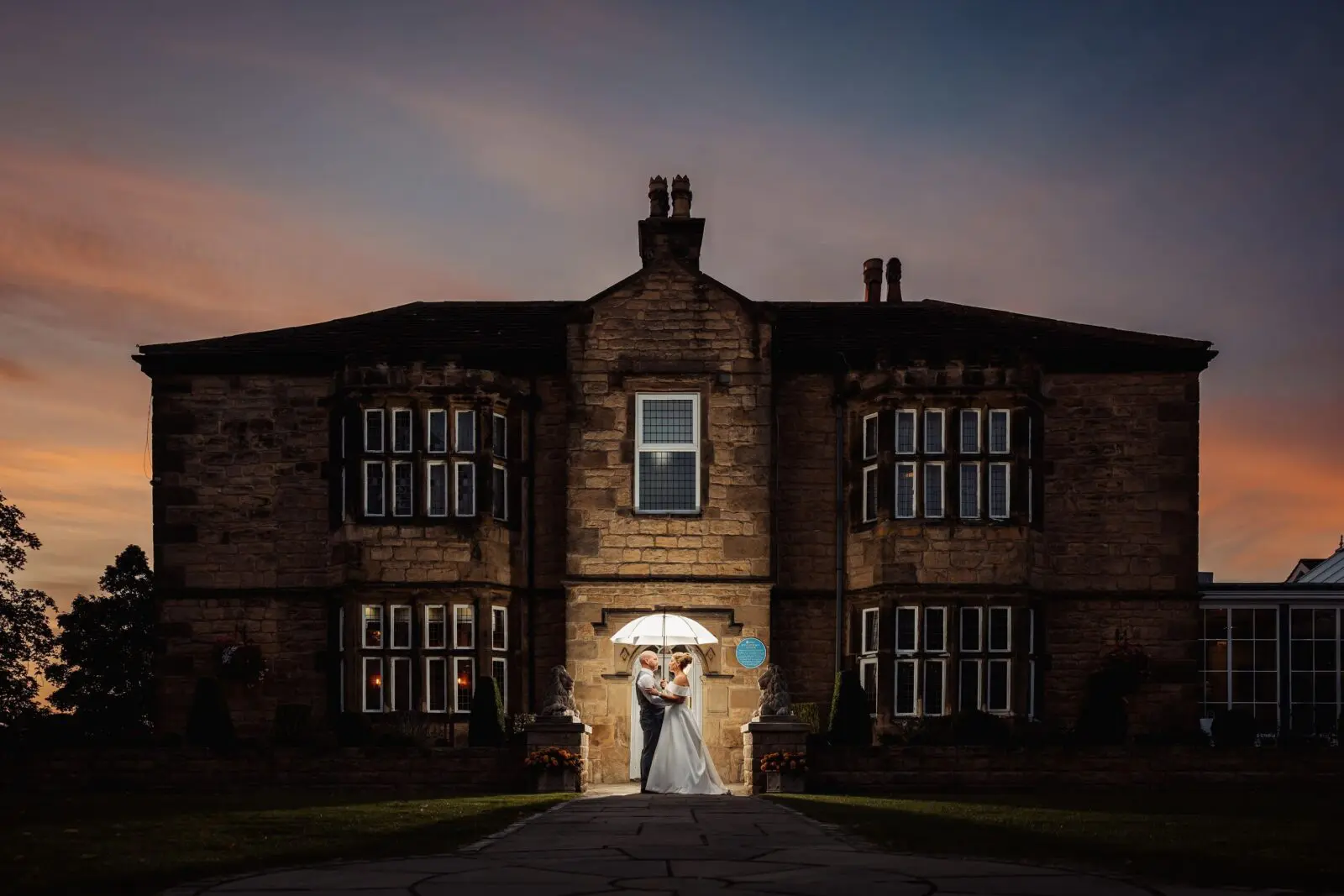 A couple stands under a white umbrella in front of a large stone building at dusk.