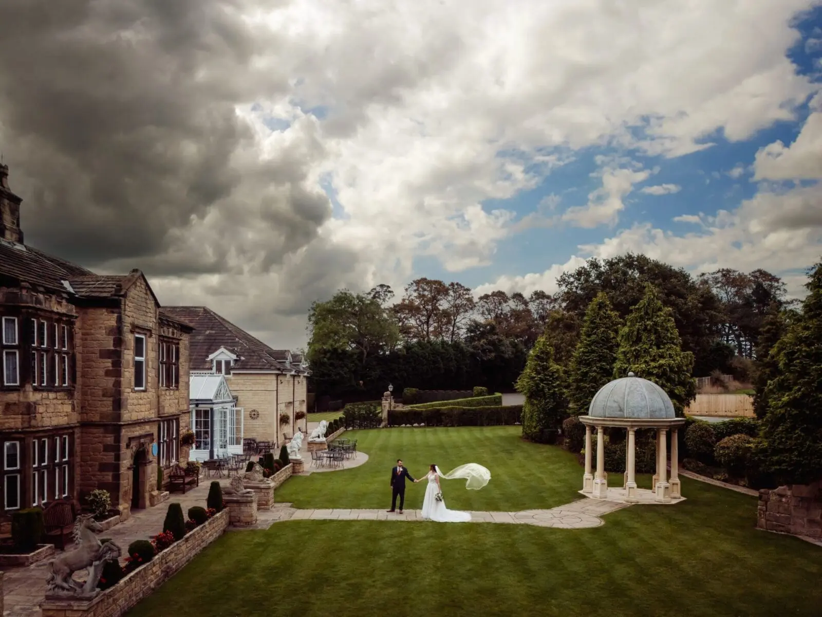 A couple in wedding attire walks across a spacious garden with manicured lawns, a large house on the left, and a white gazebo on the right under a cloudy sky.
