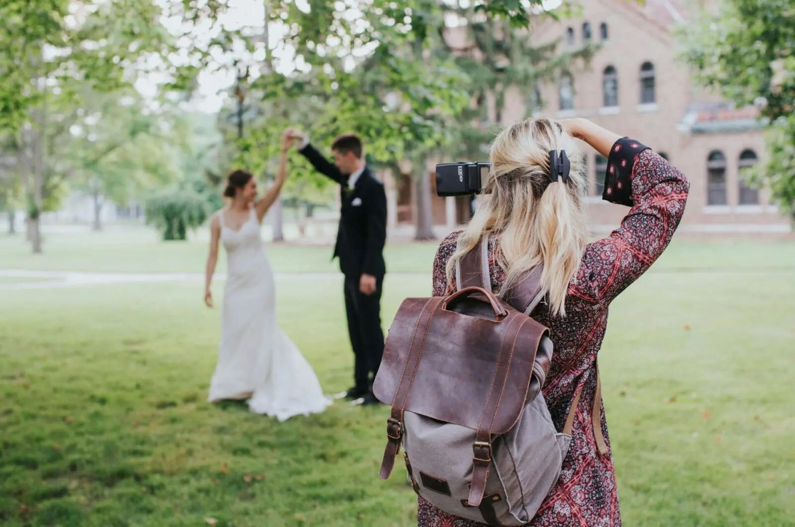 A wedding photographer takes an photo of a bride and groom on the grass under a tree