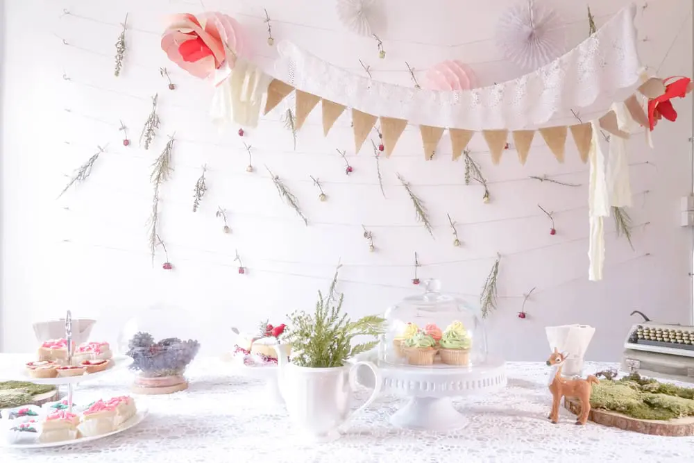 A decorated table showcases cupcakes, cookies, and floral arrangements alongside a vintage typewriter and deer figurine, perfect for wedding planning. A paper garland and dried flowers elegantly adorn the wall backdrop, creating a charming atmosphere ideal for any wedding UK setting.