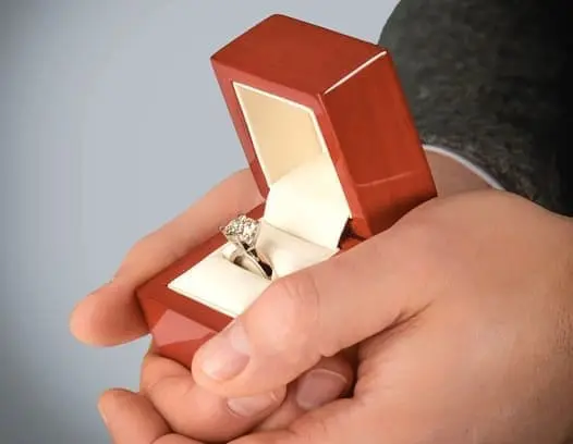 A person holds an open red ring box containing a diamond ring.