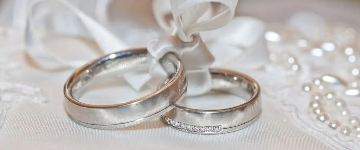 Two platinum and gold wedding rings sit on a white lace surface, tied together with a white satin ribbon. One ring has a row of small diamonds.