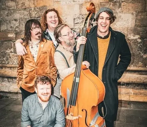 Five people, three standing and two kneeling, pose with a double bass against a stone wall, smiling and laughing.