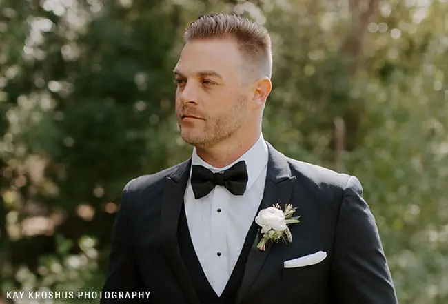 A man in a tuxedo with a bow tie and boutonniere stands outdoors in a forested area.