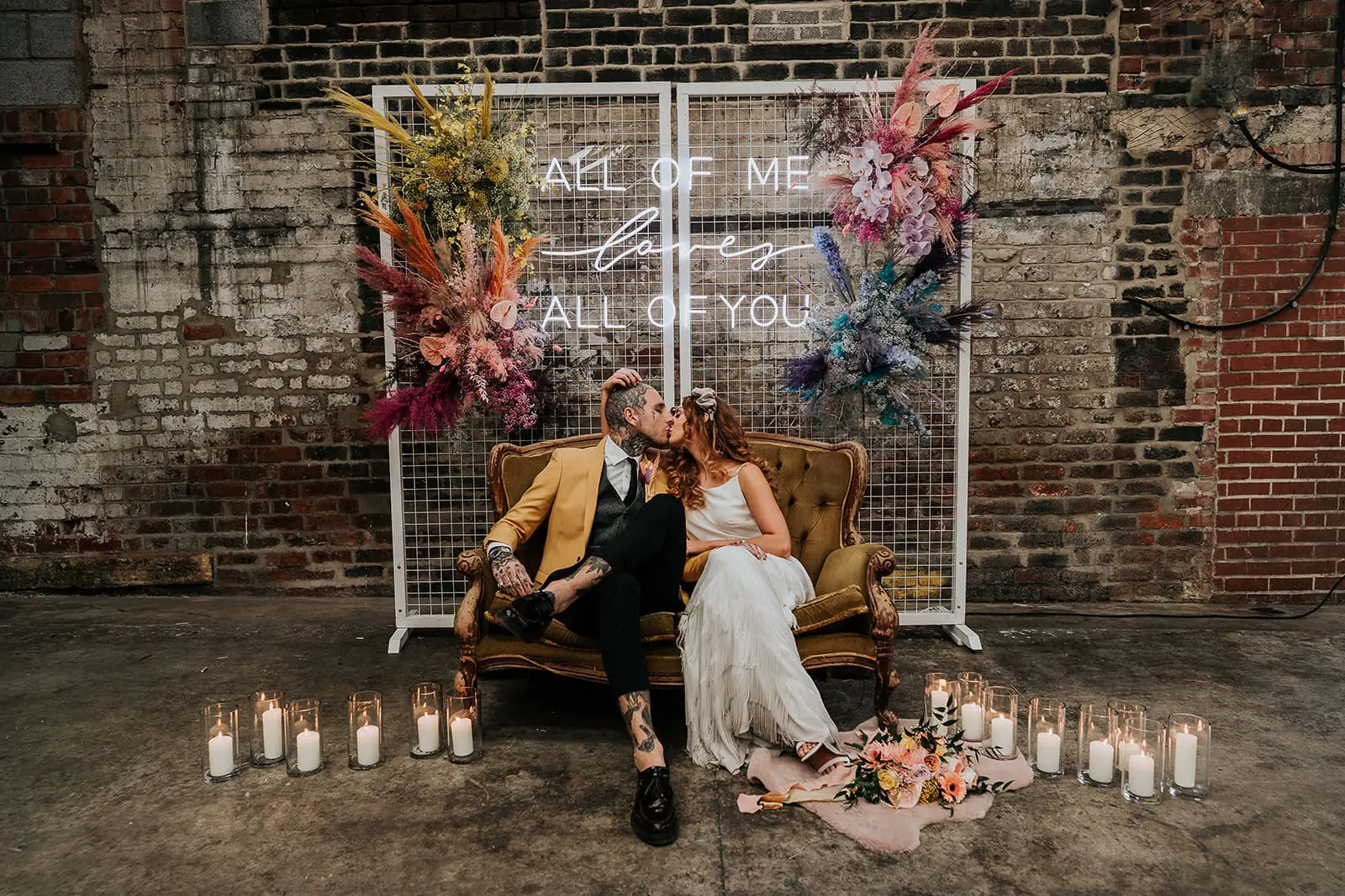 A couple in wedding attire sits on a vintage couch in an industrial-style venue. Behind them is a sign reading "All of me loves all of you," with flowers and candles adorning the scene.