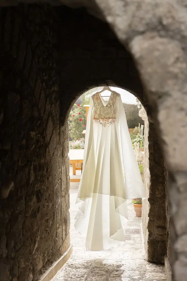 A white wedding dress hangs in a stone archway, with an outdoor garden visible in the background.