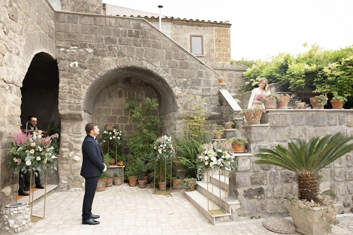 A man in a suit stands on a stone patio with floral arrangements, looking up at a woman in a dress on stairs. Another man holds a guitar nearby.
