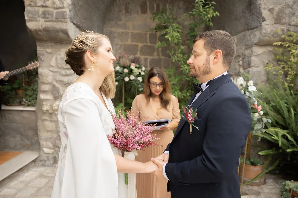 A bride and groom hold hands in an outdoor ceremony, with a person standing between them, reading from a book. Background features stone walls and greenery.
