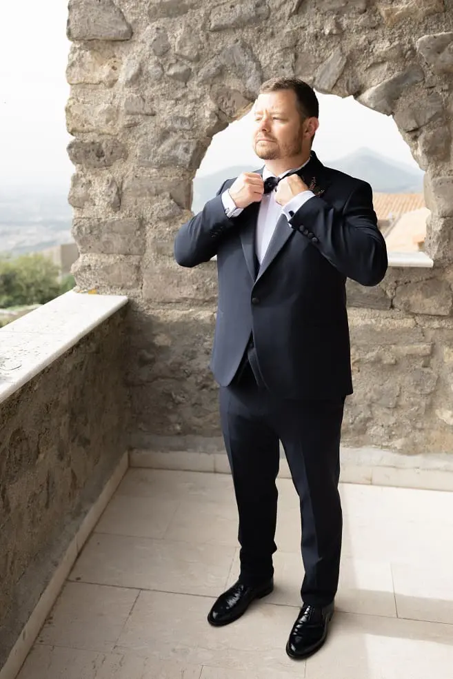A man in a suit adjusts his tie on a stone balcony with a scenic view in the background.