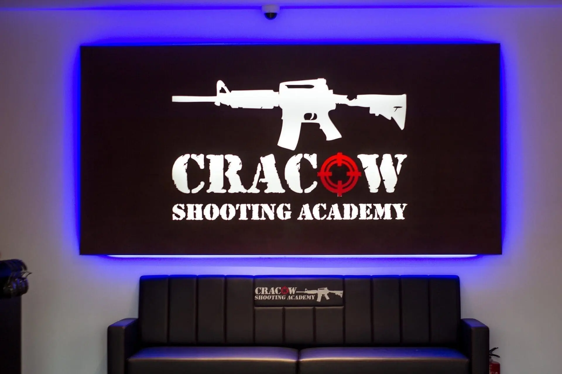A sign for Cracow Shooting Academy with a rifle graphic is illuminated in blue above a black sofa.