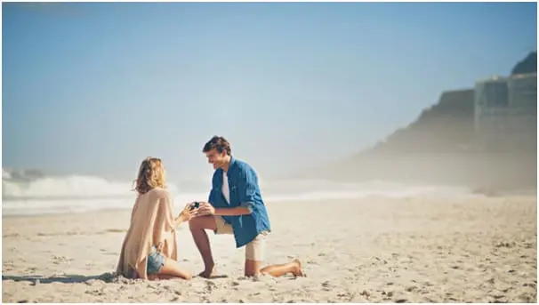 A man is kneeling on a sandy beach, holding a ring, proposing to a woman sitting in front of him. The ocean and a hazy sky are in the background.